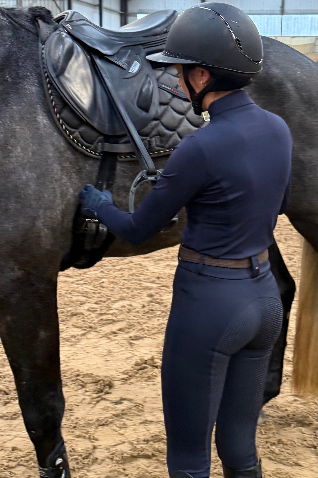 Person in equestrian attire adjusting a horse's saddle in an indoor arena.