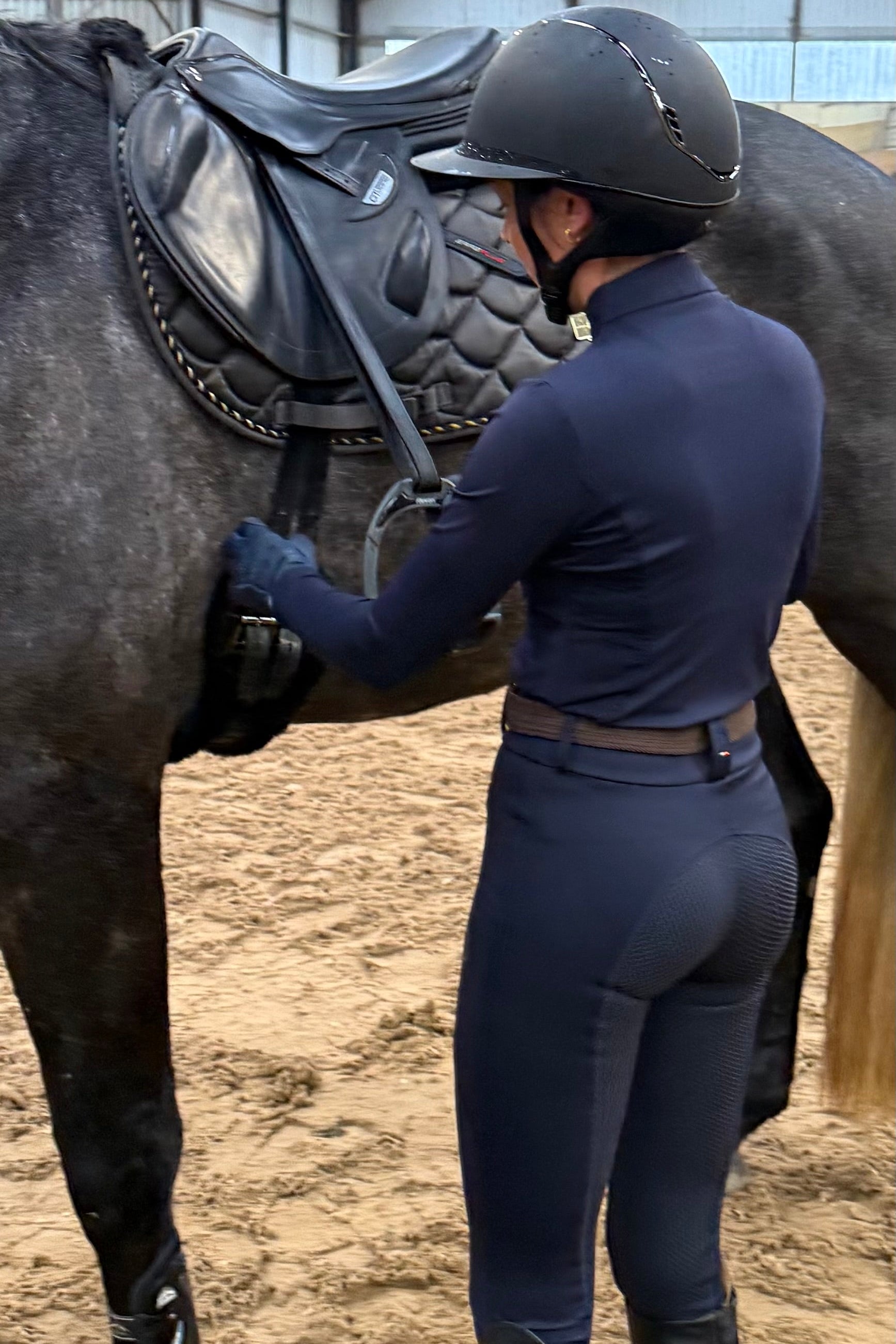 Person in equestrian attire adjusting a horse's saddle in an indoor arena.