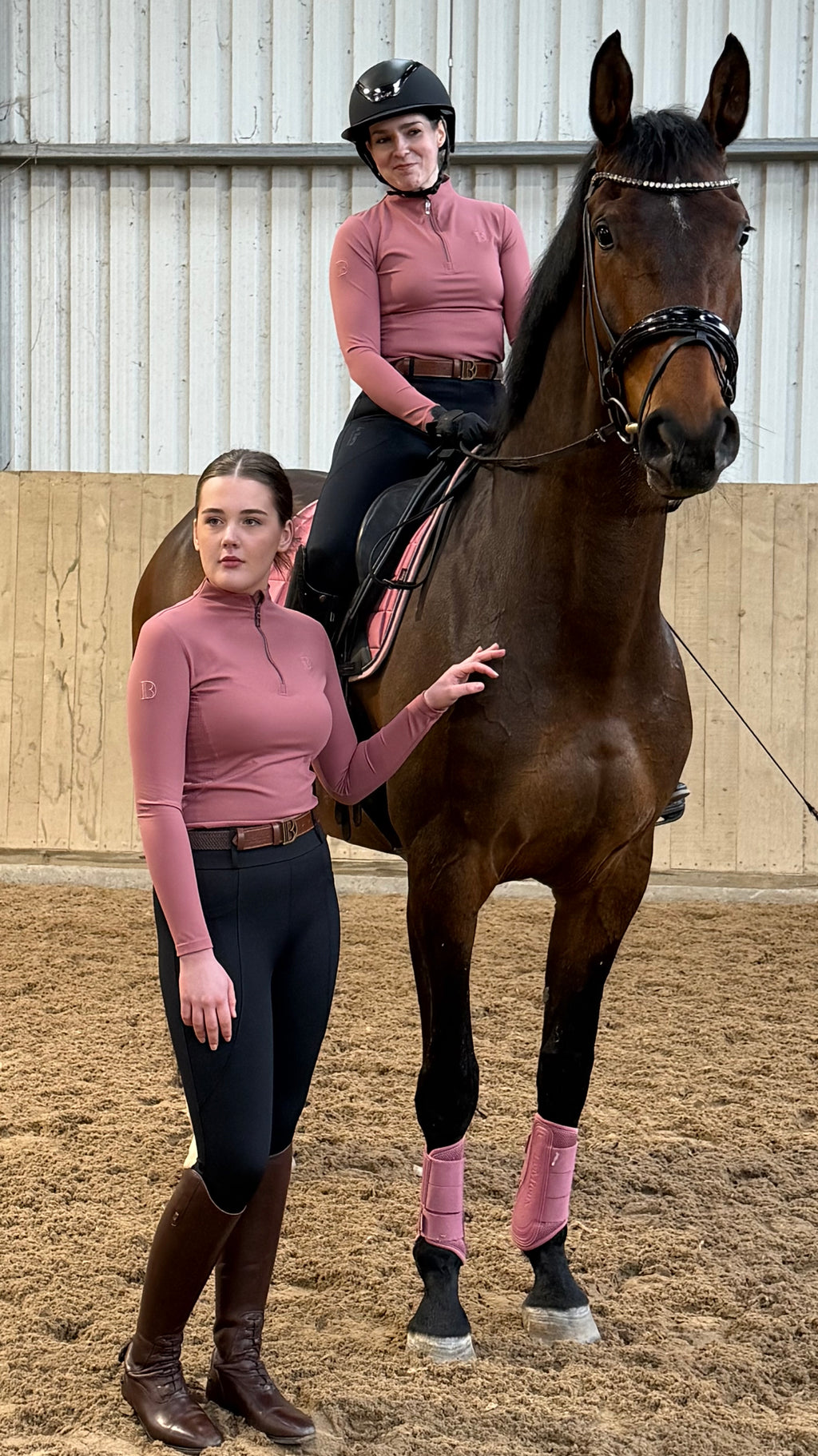 Two people in equestrian attire standing with a brown horse in an indoor arena.