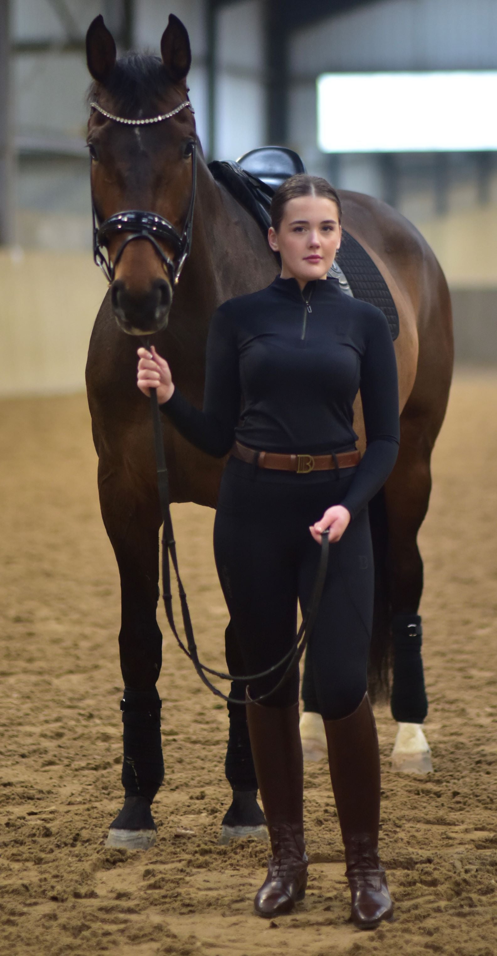 Woman in equestrian attire standing next to a horse in an indoor arena.
