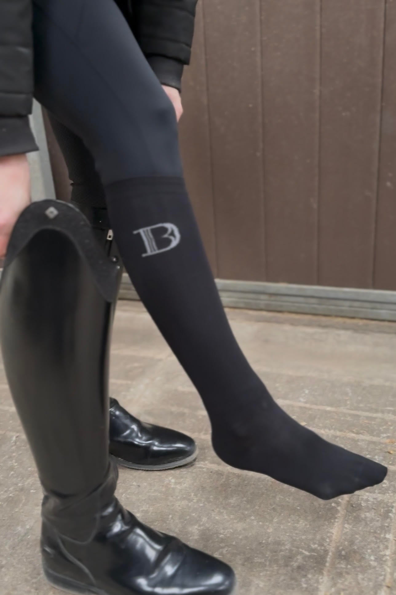 Person wearing black riding boots and socks with a logo on a concrete floor.